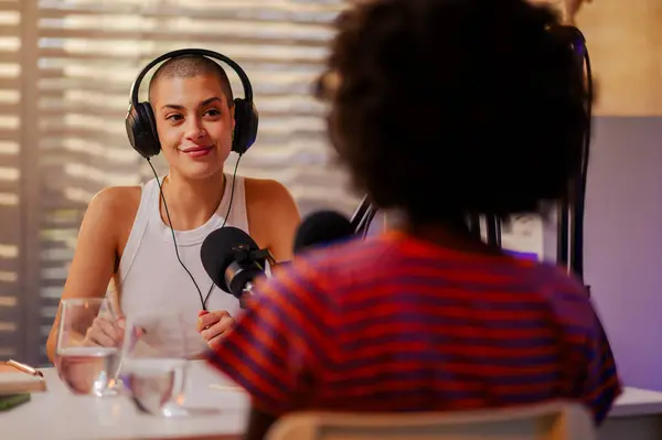 stock image Two multiracial girls are sitting in a small recording studio at home with headphones on and recording video content for the internet. Two podcasters are recording a vlog during a podcast episode.