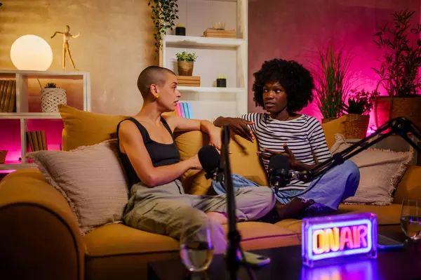 stock image Young multiracial women smiling while recording an audio broadcast in a home studio. Cheerful diverse content creators co-hosting an internet podcast. Bald caucasian and african american woman.