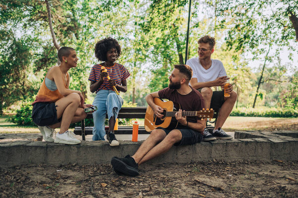 A group of multicultural gen z friends sitting at city park with guitar and having fun time together. A young man is playing a guitar to his friends. Young people listening to their friend with guitar