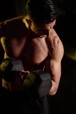 Spor salonunda antreman yaparken kaslı üstsüz adam dambılı kaldırıyor.