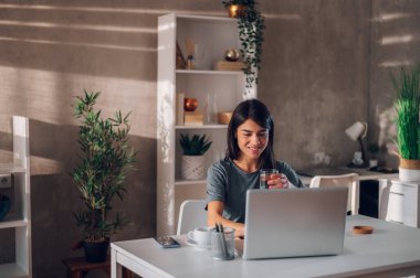Koyu renk saçlı bir iş kadınının elinde bir bardak su tutarken evinde oturup laptopuyla çalışmasının geniş açılı görüntüsü. Mücadeleci ve mutlu görünüyorsun. Uzaktan çalışma konsepti. Boşluğu kopyala.