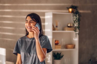 Mutlu genç bir iş kadınının portresi. Güneşli evinde durmuş cep telefonuyla konuşuyor ve gülüyor. Bir arkadaşla telefon görüşmesi yapmak için işe kısa bir ara verdim. Boşluğu kopyala.