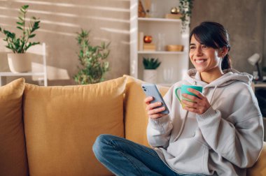 Evdeki kanepede oturup kahve içerken akıllı telefon kullanan genç bir kadın. İletişim, bağlantı, mobil uygulamalar, teknoloji, öğrenme, sohbet, yaşam tarzı kavramı.