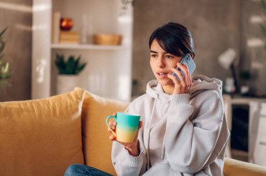 Evdeki bir kanepede oturan bir kadının portresi ve arkadaşlarıyla ya da ailesiyle akıllı bir telefonla konuşması. Mutlu bir kadın cep telefonu kullanıp koltukta otururken kahve içiyor. Boşluğu kopyala.