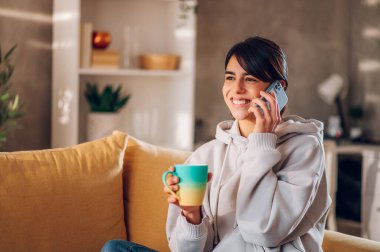 Kanepede rahat bir şekilde oturan, akıllı telefonla konuşan ve kahve içen bir kadın. Gülümseyen genç bayan evde dinlenirken arkadaşlarıyla ve ailesiyle konuşmaktan zevk alıyor. Boşluğu kopyala.
