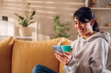 Kanepede rahat bir şekilde oturan bir kadın kahve içerken ve sosyal medyada akıllı telefondan iyi haberler verirken. Gülümseyen genç bayan cep telefonu uygulamasında hafta sonu yemeği siparişi veriyor..