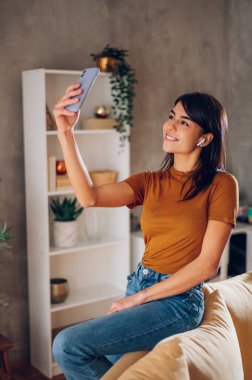Güzel bir kadın evde dinlenirken akıllı telefonuyla selfie çekiyor. Kendi fotoğrafını çekmek için cep telefonu kullanan gülümseyen genç bir kadın. Boşluğu kopyala.