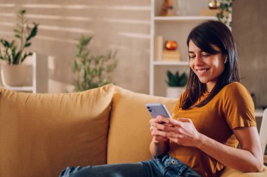 Evdeki kanepede yatarken akıllı telefon kullanan genç bir kadın. İletişim, bağlantı, mobil uygulamalar, teknoloji, öğrenme, sohbet, yaşam tarzı konsepti. Boşluğu kopyala.