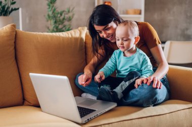 Memnuniyetle anne ya da bebek bakıcısı gülümseyen ve kucak kucak açan çocuk evde kanepede dinlenirken ve dizüstü bilgisayar kullanırken. Mutlu aile hayatı, çizgi film izlemek. Boşluğu kopyala.