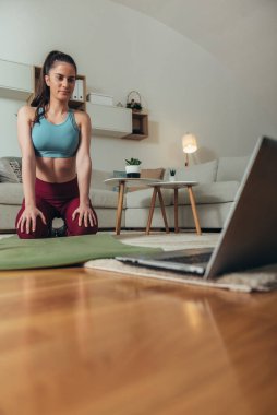 Genç ve çekici bir kadın evde esniyor ve online fitness eğitimi için dizüstü bilgisayar kullanıyor.