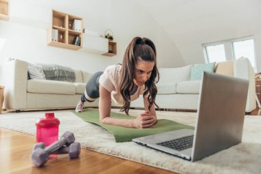 Tahta bir pozisyonda çalışırken ve online fitness sınıfı için dizüstü bilgisayar kullanırken evde çalışan çekici genç bir kadın.
