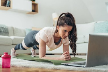 İnternet dersini kullanırken evde tahta egzersizleri yapan genç bir kadının fotoğrafı.