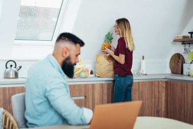 Mutlu genç bir çiftin portresi laptoplarını kullanırken evlerindeki mutfak malzemelerini boşaltıyor.