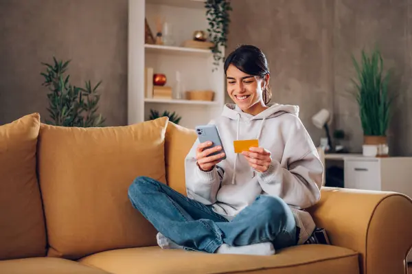 Evdeki bir kanepede otururken elinde akıllı telefon ve kredi kartı olan güzel beyaz bir kadın. Gülümseyen genç bir kadın internetten alışveriş yapıyor ve mobil uygulamadan ürün sipariş ediyor..