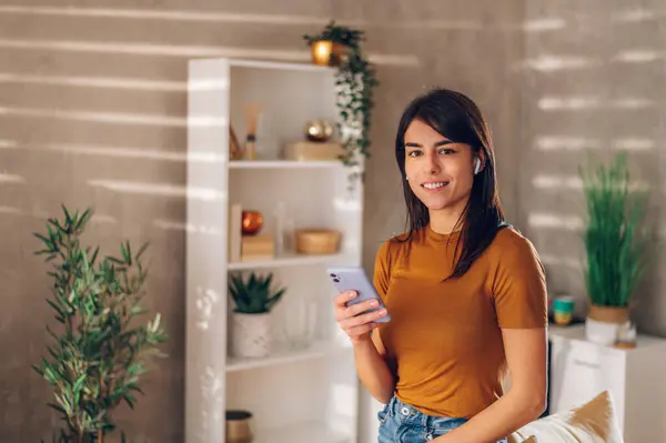 Stock image Portrait of a woman relaxing at home and holding smartphone while having airpods in her ears. Beautiful young lady chatting and using social media on her mobile phone. Copy space.