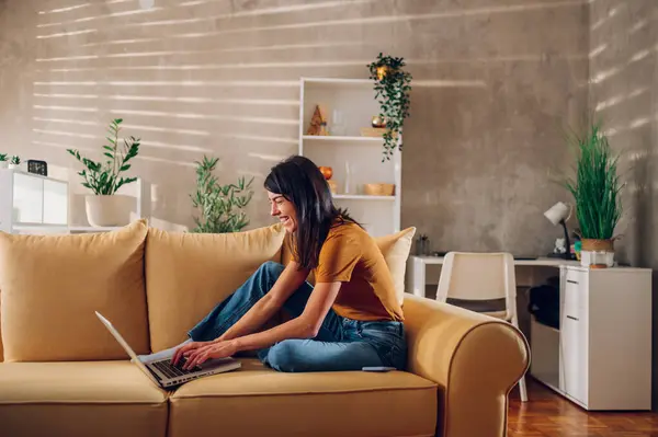 Evde kanepede otururken dizüstü bilgisayar kullanan gülümseyen beyaz bir kadının yan portresi. Mutlu kadın alışverişleri ya da sosyal ağda online sohbetler bilgisayar projesinde çalışan serbest çalışan biri. Boşluğu kopyala.