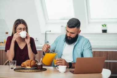 Genç ve çekici bir çift evde kahvaltı yaparken dizüstü bilgisayar kullanıyor ve kahve içiyor.