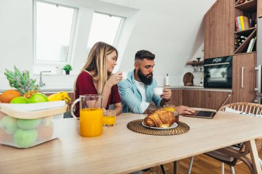 Genç ve mutlu bir çiftin evde kahvaltı ederken dizüstü bilgisayar kullanması ve kahve içmesi.