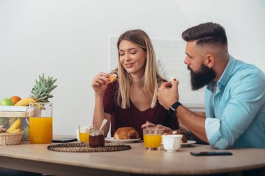Evde, mutfakta kahvaltı yapan genç bir çiftin fotoğrafı.