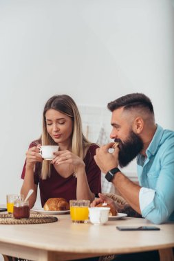 Genç bir çift evdeki mutfakta kahvaltı ederken kahve içiyor.