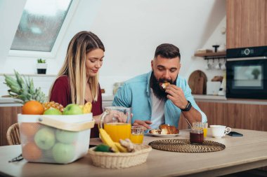 Çift evde kahvaltı yaparken portakal suyu içip kruvasan yiyor.