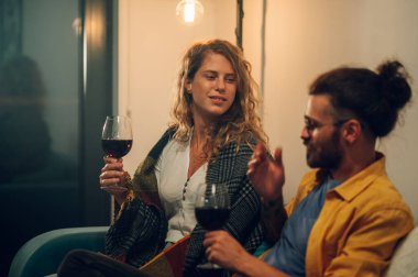 Sarışın, genç, kıvırcık bir kızın, erkek arkadaşıyla sıcak bir kış kulübesinde otururken yan görünüm portresi. Elinde bir kadeh kırmızı şarapla onu dinlerken. Birlikte kaliteli zaman geçirmek..