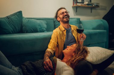 Kız arkadaşı kucağında yatarken genç adamın yere oturduğu yakın çekim. Elinde şarap kadehi olan ve gözleri kapalı gülen bir adama odaklan. Boşluğu kopyala..