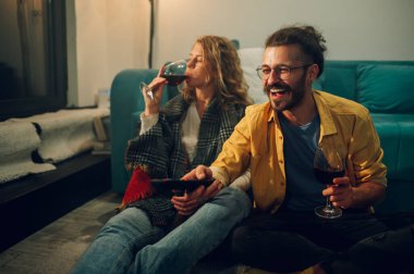 Yerde oturup uzaktan kumandayla kanal değiştiren mutlu bir gencin yan görüntüsü. Kız arkadaşıyla hafta sonunun tadını çıkarırken şarap yudumluyor ve sıcak bir kulübede vakit geçiriyor..