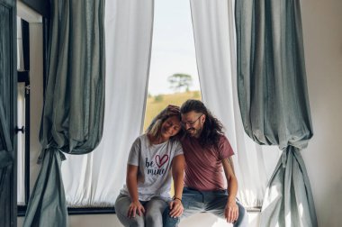 Genç çift evde birlikte eğleniyor. Kadın ve erkek yatak odasındaki pencerenin yanında oturup sarılıyorlar. Yetişkinler arası ilişki. Boşluğu kopyala.