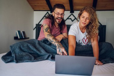 Güzel mutlu genç çift bir hafta sonu uyandıktan sonra birlikte gülerken dizüstü bilgisayar kullanıyor ve internette internette geziniyor. Yatakta planlama günü. Haber okuyor, e-postayı yeniden oynatıyor.