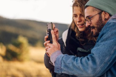 Sıcak kıyafetler giymiş birkaç turist çimenli bir dağ sırtında yürüyorlar ve bir manzaranın fotoğrafını çekmek için akıllı telefon kullanıyorlar. Doğada yürüyüş yapan gençler. Cep telefonuna odaklan. Boşluğu kopyala.