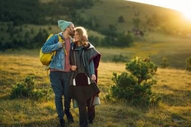 Sıcak kıyafetler giymiş romantik turistler gün batımında çimenli bir dağ sırtında yürürken sarılıp öpüşüyorlar. Doğada yürüyüş yapan gençler. Boşluğu kopyala.