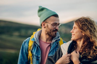 Gün batımında çimenli bir dağ sırtında yürüyen kalın kıyafetler giymiş romantik bir çift turistin portresi. Tarz sahibi bir adam ve birbirine aşık bir kadın. Boşluğu kopyala.