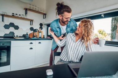 Mutfak masasında dizüstü bilgisayarda çalışan mutlu bir kadın erkek arkadaşları ona evde sarılırken. Elektronik cihazlar kullanarak modern çağlar. Elinde akıllı telefon ve bir fincan kahve tutan bir adam. Boşluğu kopyala.