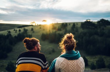 Mutlu bir çiftin sabah kahvesinin tadını çıkarırken ve arka planda güzel bir doğa ve gün doğumunda sıcak bir ışıkla balkonda dikilirken başka yere bakarken görüntüsü..