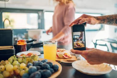 Mutfakta kahvaltıyı çekerken akıllı telefon tutan bir adamın yakın çekimi. Karı koca hafta sonu evde yemek hazırlıyor ve eğleniyorlar..