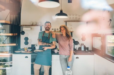 Sabahları sıcacık bir mutfakta yemek yapmaktan zevk alan genç bir çiftin portresi. Kadın kameraya bakarken erkeğine sarılıyor. Sevgi dolu çift yemek yaparken eğleniyor. Boşluğu kopyala.