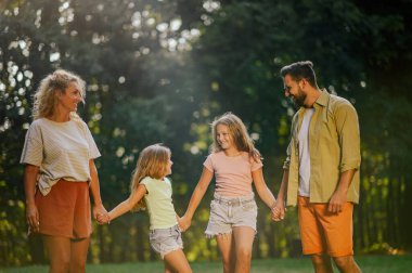 Neşeli bir ebeveyn kızlarıyla el ele tutuşuyor ve hafta sonları güneşli bir yaz gününde doğa yürüyüşü yapıyor. Dört kişilik mutlu bir aile hafta sonları doğada vakit geçirir. Boşluğu kopyala.