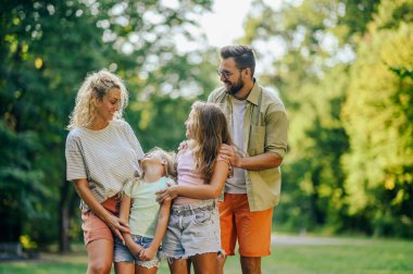 Neşeli bir aile ormanın yakınındaki çayırda duruyor ve birlikte iyi vakit geçiriyorlar. Genç bir aile güneşli bir yaz gününü doğada geçiriyor. Anne ve baba kızlarıyla birlikte doğada duruyorlar..