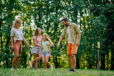 Hafta sonları güneşli yaz günlerinde doğada kızlarıyla eğlenen ebeveynlerin portresi. Ormanın yakınındaki bir çayırda yürürken anne babalar onlara bakarken oynayan kızların portresi. Boşluğu kopyala.