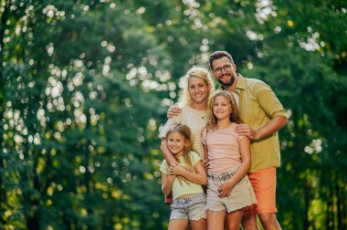 Doğadaki ormanın yakınındaki kameraya sarılıp gülümseyen mutlu bir ailenin portresi. Genç bir aile güneşli bir yaz gününde doğada birlikte sarılıp eğleniyor. Doğada aile resmi.