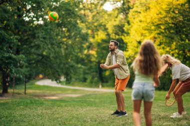 Doğada voleybol oynarken topa vuran genç bir babanın portresi. Onun yanında badminton oynayan anne ve kızı var. Sportif bir aile spor gününü doğada birlikte geçiriyor..