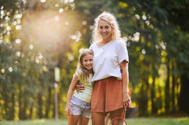 Doğada mahsur kalmış ve kameraya gülümserken Badminton raketlerini kucaklayan mutlu anne ve çocuğun portresi. Aktif, sportif bir aile günü. Boşluğu kopyala.