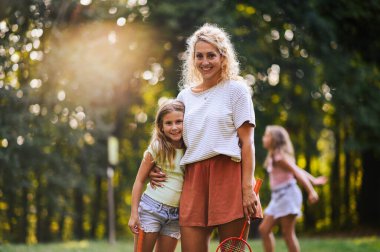 Güneşli bir yaz gününde doğada badminton raketleri tutarken kızını kucaklayan sevgi dolu bir annenin portresi. Mutlu anne, doğada durup kameraya gülümserken kızını kucaklıyor..
