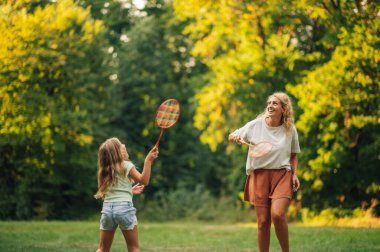 Doğada eğlenen mutlu bir anne ve kız raket tutarken ve topa vururken badminton oynuyorlar. Doğada badminton oynayan sportif bir ailenin portresi. Etkin tatil.