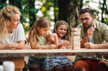 Neşeli bir kız, doğada ailesi ve kız kardeşiyle eğlenceli bilişsel Jenga oyunu oynarken hamlesini yapıyor. Eğlenceli aile portresi piknikte masa oyunu günü..