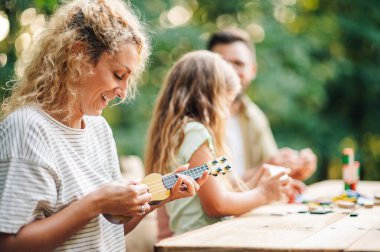 Neşeli, oyuncu bir annenin doğada ailesiyle birlikte oturduğu ve ukulele oyuncağıyla müzik yaptığı profil görüntüsü. Mutlu anne bulanık arka planda oynayan ailesini eğlendiriyor..