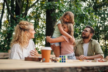 Sevgi dolu kız kardeşler doğadaki piknik masasında dururlar ve ebeveynleri onlara gülümserken kucaklaşırlar. Mutlu çocuklar sevgi gösteriyor. Kız kardeşler birbirlerine sarılırken aileleri onlara gülümsüyor..