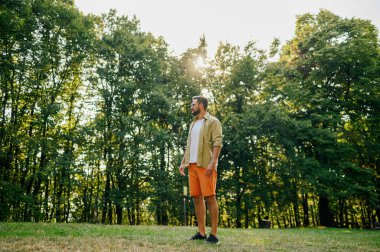 Ormanın yakınındaki bir çayırda duran ve güneşli bir yaz gününün tadını çıkaran genç bir adam. Bir adam haftasonunu doğada geçiriyor ve doğal güzellikleri ulusal parkta keşfediyor. Boşluğu kopyala.