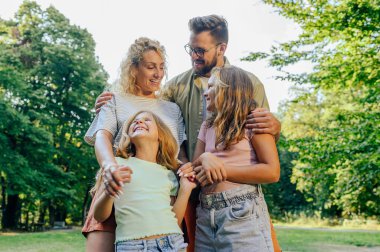 Doğada durup kucaklaşan kızlarıyla neşeli bir ebeveynin portresi. Genç bir anne ve baba kızlarıyla ormanın yakınında durup onlara sarılıyorlar. Aile içinde aile.